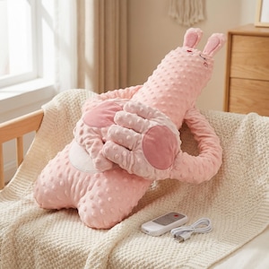 Peut inclure: Un oreiller rose en forme de lapin avec une surface texturée et des éléments en forme de mains attachés. L'oreiller a un visage souriant et de longues oreilles. Une télécommande et un câble de chargement sont visibles.