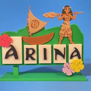 Puede incluir: Un letrero decorativo verde y blanco con el nombre "ARINA" en letras negras. Presenta un barco, un personaje con una paleta y acentos florales. El letrero est&aacute; sobre una base verde sobre un fondo azul.