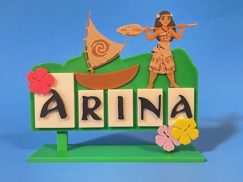 Puede incluir: Un letrero decorativo verde y blanco con el nombre "ARINA" en letras negras. Presenta un barco, un personaje con una paleta y acentos florales. El letrero est&aacute; sobre una base verde sobre un fondo azul.