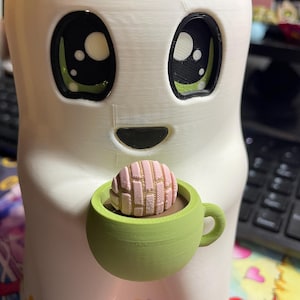 May include: A white 3D-printed ghost figurine with large black and green eyes, holding a small light green mug containing a brown liquid and a pink concha bread roll with a textured grid pattern on top, featuring a smooth matte finish.