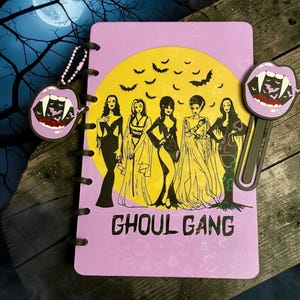 May include: A light purple notebook with a yellow moon graphic featuring five female figures and the text GHOUL GANG. It includes a matching purple vampire mouth charm on a beaded chain and a purple vampire mouth bookmark with a long stem.