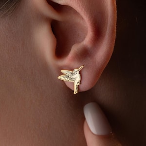 May include: A close-up of a gold-colored hummingbird stud earring on a person's ear. The earring is detailed with a small, sparkling accent. The person's skin is fair, and their fingernails are painted white. The earring is a jewelry item.