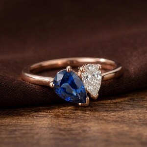May include: Rose gold ring featuring two pear-shaped gemstones, one a deep blue sapphire and the other a clear diamond. The ring is set against a dark brown fabric and a wooden surface, showcasing the jewelry's elegant design.