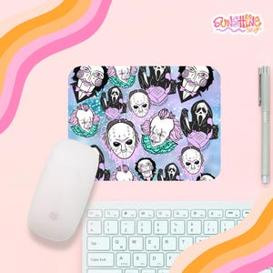 Puede incluir: Una alfombrilla de ratón rectangular con un patrón repetido de personajes de películas de terror, incluyendo a Jigsaw, Pennywise y Ghostface. La alfombrilla tiene un fondo azul claro y morado. También se ven un ratón, un teclado y un bolígrafo blancos.