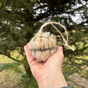 May include: A beige, spherical ornament with a grid pattern, suspended by a natural twine loop. The ornament is held in a hand against a backdrop of green evergreen branches, suggesting a rustic, natural theme.