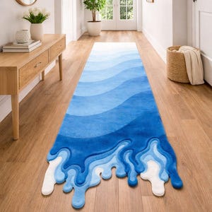 May include: A blue and white runner rug with a unique wave design. The rug features varying shades of blue, creating a gradient effect. The bottom edge has a stylized, dripping wave pattern. The rug is placed on a wooden floor.
