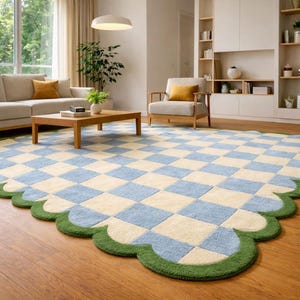 May include: A large, checkered rug with a scalloped edge in a living room setting. The rug features a pattern of light blue and cream-colored squares, bordered by a green scalloped edge. The room includes a beige sofa, wooden coffee table, and armchair.