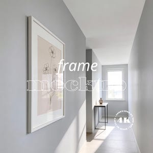 Puede incluir: Una impresión artística enmarcada en blanco con un dibujo lineal floral cuelga de una pared gris en un pasillo. El texto "frame mockup" está superpuesto en la imagen. Se ve una pequeña mesa con un jarrón.