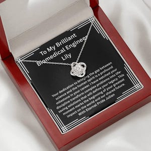 May include: A silver-toned necklace with a central knot pendant featuring a clear stone, presented in a mahogany-colored box. The card inside reads: To My Brilliant Biomedical Engineer Lily, followed by a message about bridging science and spirit.