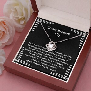 May include: A silver-toned necklace with a central love knot pendant featuring a clear round stone, presented in a dark wooden box. The black insert displays white text addressed to Lily, praising analytical precision and achievement, signed by Julian.