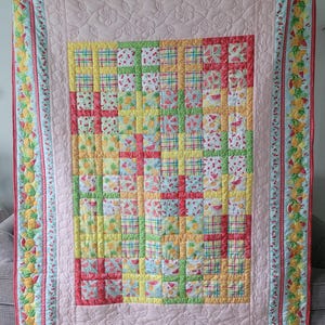 May include: A colorful quilt with a patchwork design featuring squares of fruit patterns, including watermelons, cherries, and pineapples. The quilt has a light pink background with a floral border and a blue trim with fruit designs. The quilt is approximately 70 inches by 90 inches.