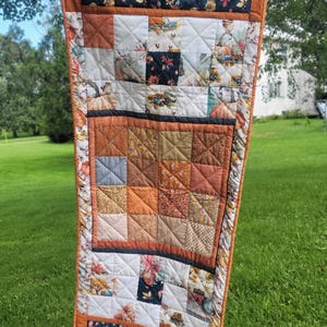 May include: A rectangular quilted table runner featuring a patchwork design with autumn-themed fabric prints. The quilt displays squares of orange, brown, and cream, accented by dark navy panels with floral and truck motifs, all finished with an orange border.