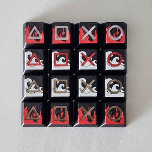 Puede incluir: Un conjunto de dieciséis teclas negras con símbolos de juego, incluyendo triángulos, cuadrados, cruces y círculos. Cada tecla presenta un diseño de llama roja en la parte superior, dispuestas en una cuadrícula de 4x4.