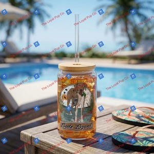 May include: A clear glass tumbler with a bamboo lid and a glass straw, featuring a graphic of a brown and white dog standing in water with tall grass. The label includes the text Bode Byand in a script font, filled with a golden liquid.