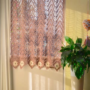 May include: A dusty rose crochet curtain featuring a repeating chevron pattern with openwork details and a scalloped bottom edge adorned with small, cream-colored floral motifs. The fabric is semi-sheer, allowing light to pass through the intricate yarn work.
