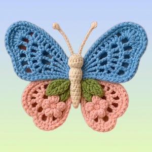 Op de afbeelding: Een gehaakte vlinder met lichtblauwe bovenvleugels met opengewerkt patroon, roze ondervleugels met roze bloemen, een crèmekleurig lijfje, twee crèmekleurige voelsprieten en twee groene blaadjes bij het midden.