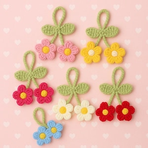 Puede incluir: Adornos florales de ganchillo en varios colores, incluyendo rosa, amarillo, rojo, blanco y azul, cada uno con un centro amarillo. Las flores están unidas a hojas y tallos verdes, creando un encantador diseño floral. El fondo es rosa claro con formas de corazón.