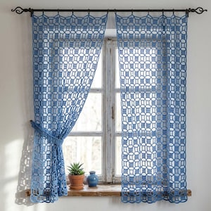 Puede incluir: Un par de cortinas de ganchillo azul con un patrón geométrico calado de círculos y cuadrados entrelazados. Las cortinas cuelgan de una barra de metal negro con anillas y un lado está recogido con un alzapaños de ganchillo a juego.