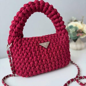 Op de afbeelding: Bordeaux gehaakte handtas met een zilveren kettingriem en handvat. De tas heeft een rechthoekige body en een driehoekig zilveren logo. De handgreep is dik en afgerond, en de kettingriem is ook bordeaux.