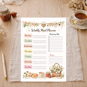 May include: A Weekly Meal Planner with floral and strawberry illustrations. The planner includes sections for each day of the week, a grocery list, and notes. A pen, tea cup, and jam jar are also visible.