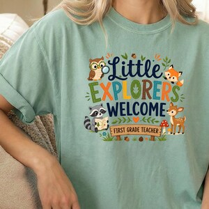 May include: A sage green short-sleeve t-shirt featuring a graphic print with the text Little Explorers Welcome First Grade Teacher. The design includes illustrations of an owl, a raccoon holding a map, a fox, and a deer among forest elements.