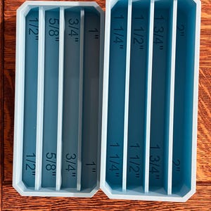 May include: Two rectangular, teal-colored resin molds with vertical dividers. The molds are labeled with inch measurements: 1/2", 5/8", 3/4", 1", 1 1/4", 1 1/2", 1 3/4", and 2". The molds are designed for crafting or creating items.