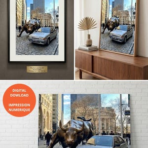 Puede incluir: Impresión enmarcada que presenta la icónica estatua del toro de Wall Street en la ciudad de Nueva York, con un coche plateado en primer plano. La imagen está ambientada contra un telón de fondo de edificios altos y un cielo azul. La impresión se titula "The Bull and the BX".