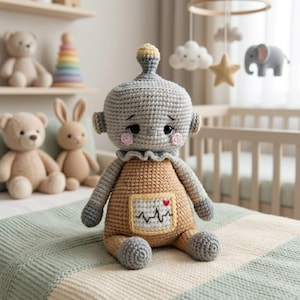 Puede incluir: Muñeca robot de ganchillo hecha a mano en gris y marrón, con mejillas rosas y un diseño de monitor cardíaco en el pecho. La muñeca está sentada sobre una manta a rayas, con otros peluches en el fondo.