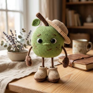 Puede incluir: Un personaje de manzana de ganchillo hecho a mano, con cuerpo verde, tallo y hoja marrones. Lleva un sombrero y botas color beige, y tiene ojos negros. La manzana está sobre una superficie de madera.