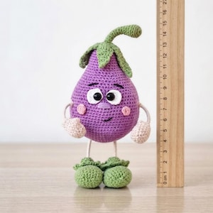 May include: A purple crochet aubergine toy with a green stem, white eyes, and pink cheeks. It has light beige arms and green feet. A wooden ruler placed next to the toy shows it stands approximately 18 cm tall from the base to the stem top.