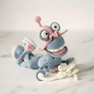 Könnte beinhalten: Handgefertigte Häkelfigur in Blau, Rosa und Weiß. Das skurrile Design zeigt große Augen, ein breites Grinsen und ausgestreckte Arme. Das Spielzeug liegt, wodurch die detaillierte Handwerkskunst und die verspielte Ästhetik zur Geltung kommen.