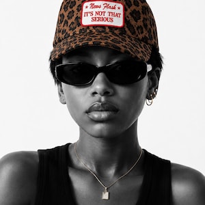 Puede incluir: Una gorra de béisbol con estampado de leopardo en marrón y negro que presenta un parche rectangular blanco con borde rojo en la parte frontal, con el texto bordado en rojo: News Flash IT'S NOT THAT SERIOUS. La gorra tiene una corona estructurada.