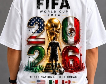 May include: A white t-shirt featuring a FIFA World Cup 2026 graphic on the back. The design includes the trophy, the number 2026 with flag patterns, a soccer player silhouette, and the text Three Nations. One Dream. United States - Mexico - Canada.