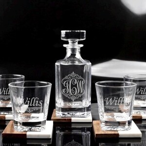 May include: A clear glass decanter with a square base and a stopper, featuring an etched monogram of the letters JCW. Four matching clear glass tumblers are included, each engraved with the name Willis. Each piece rests on a two-tone coaster.