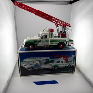 May include: A white and green Hess toy fire truck with a long, red, extendable ladder mounted on the back. The truck features the word Hess on the side and the word Gasoline on the rear fender. It is displayed on top of its original cardboard box.