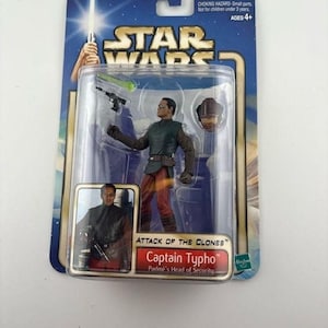 May include: A Star Wars action figure of Captain Typho from "Attack of the Clones" in a sealed package. The figure is wearing a green and brown uniform with red pants. The package includes a warning about small parts and is for ages 4+.