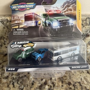 May include: A Micro Machines toy set featuring a green utility truck, a blue Warlander, and a silver bus. The packaging includes the text "Micro Machines," "Chase Rare," and a warning about small parts. The set is labeled "Micro City" and "Series 2."