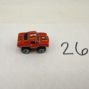 May include: A small red toy car featuring a white number 1 on the roof, black and yellow racing stripes along the top, and black wheels with white rims. The number 26 is written in black ink on the white surface next to the miniature vehicle.