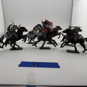 May include: A collection of miniature plastic cavalry figures on horses, painted in various colors including brown, grey, and black. The figures wear uniforms and hats, with some holding weapons. One figure carries a flag with text. A blue rectangular tag is present.
