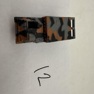May include: A toy military vehicle with a camouflage pattern in shades of brown, gray, and black. The vehicle has a rectangular shape with detailed features, including windows and a rear section with vertical slots. The number "12" is written in black ink on the white surface.