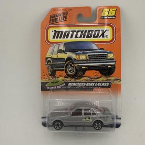 May include: A Matchbox Mercedes-Benz E-Class toy car in a clear plastic package. The car is silver with black wheels and red interior. The packaging is orange and features the Matchbox logo and the text "Guaranteed for Life".