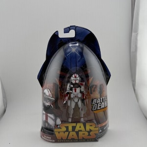 May include: A Star Wars action figure of a Clone Commander in a clear plastic package. The figure is white with red accents and is labeled "Revenge of the Sith". The packaging has a blue top and the words "Battle Gear".