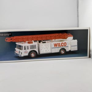May include: A white toy fire truck with a red ladder and the word "WILCO" printed on the side. The fire truck has red wheels and is displayed inside a cardboard box.