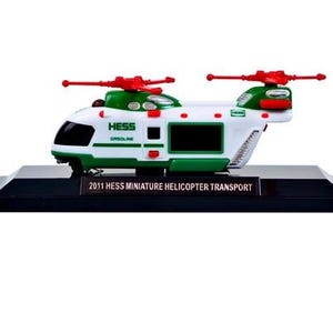 May include: A white and green miniature helicopter toy with red propellers. The toy has the word "HESS" printed on the side and sits on a black base with the text "2011 HESS MINIATURE HELICOPTER TRANSPORT".