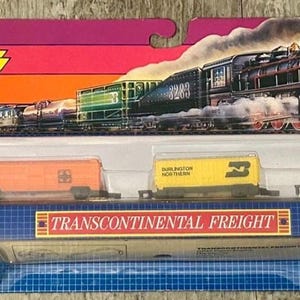 May include: A Micro Machines Transcontinental Freight train set in original packaging. The set includes a red and white locomotive, an orange tanker, a yellow Burlington Northern car, a green car, and a red caboose, with 10 tracks included.