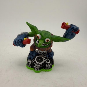 May include: A green goblin figurine with large ears, wide eyes, and a toothy grin, wearing dark blue metallic gauntlets and boots. The character holds a red dynamite stick in each hand and stands on a base featuring a black and silver gear design.