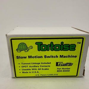 May include: A white rectangular box for a Tortoise Slow Motion Switch Machine. The yellow label features a green turtle icon, the text Tortoise, and lists features including turnout linkage, DPDT auxiliary contacts, and part number 800-6000.