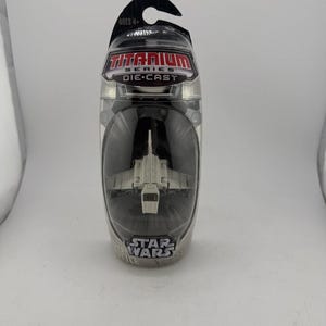 May include: A Star Wars Titanium Series die-cast model of a spacecraft, packaged in a clear plastic container. The spacecraft is white with detailed wings and body. The packaging includes the Star Wars logo and the words "Titanium Series Die-Cast."