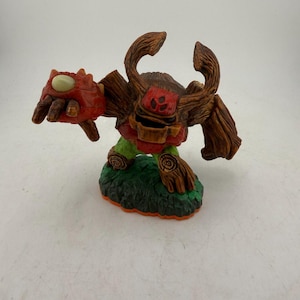 May include: A detailed figurine of a tree-like creature with a red head and arm, brown wooden body, and green and orange base. The figure has a menacing expression and is posed as if ready for action. The figure is a collectible toy.