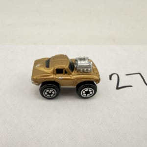 May include: A small gold-colored toy car with a prominent silver engine block protruding from the hood. The vehicle features black wheels with white star-shaped rims and black windows. The number 27 is written in black ink to the right of the car.